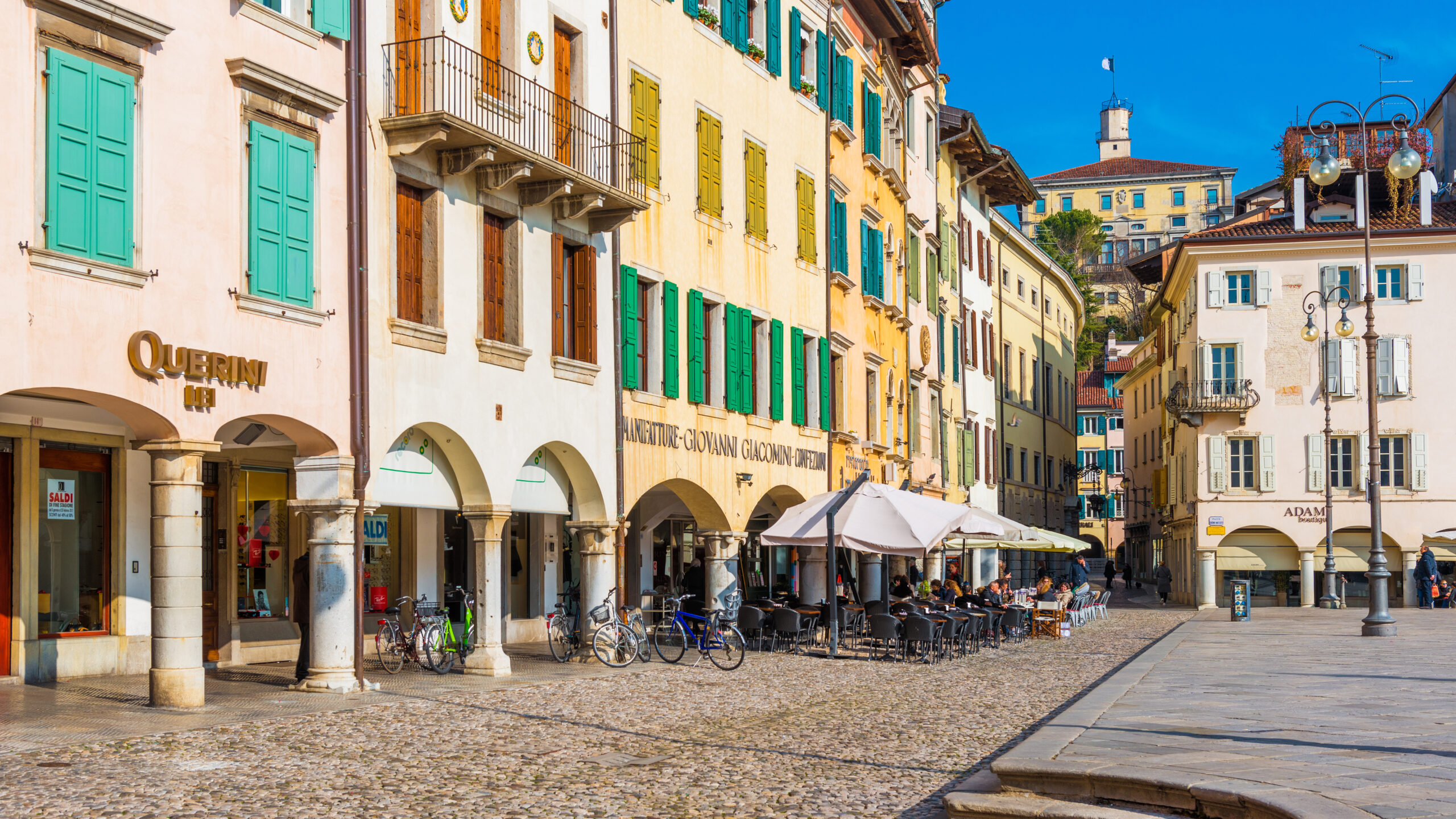 Scorcio di una piazza storica italiana con portici, negozi di prossimità e tavolini all'aperto, simbolo di rigenerazione urbana e vitalità del terziario di mercato.