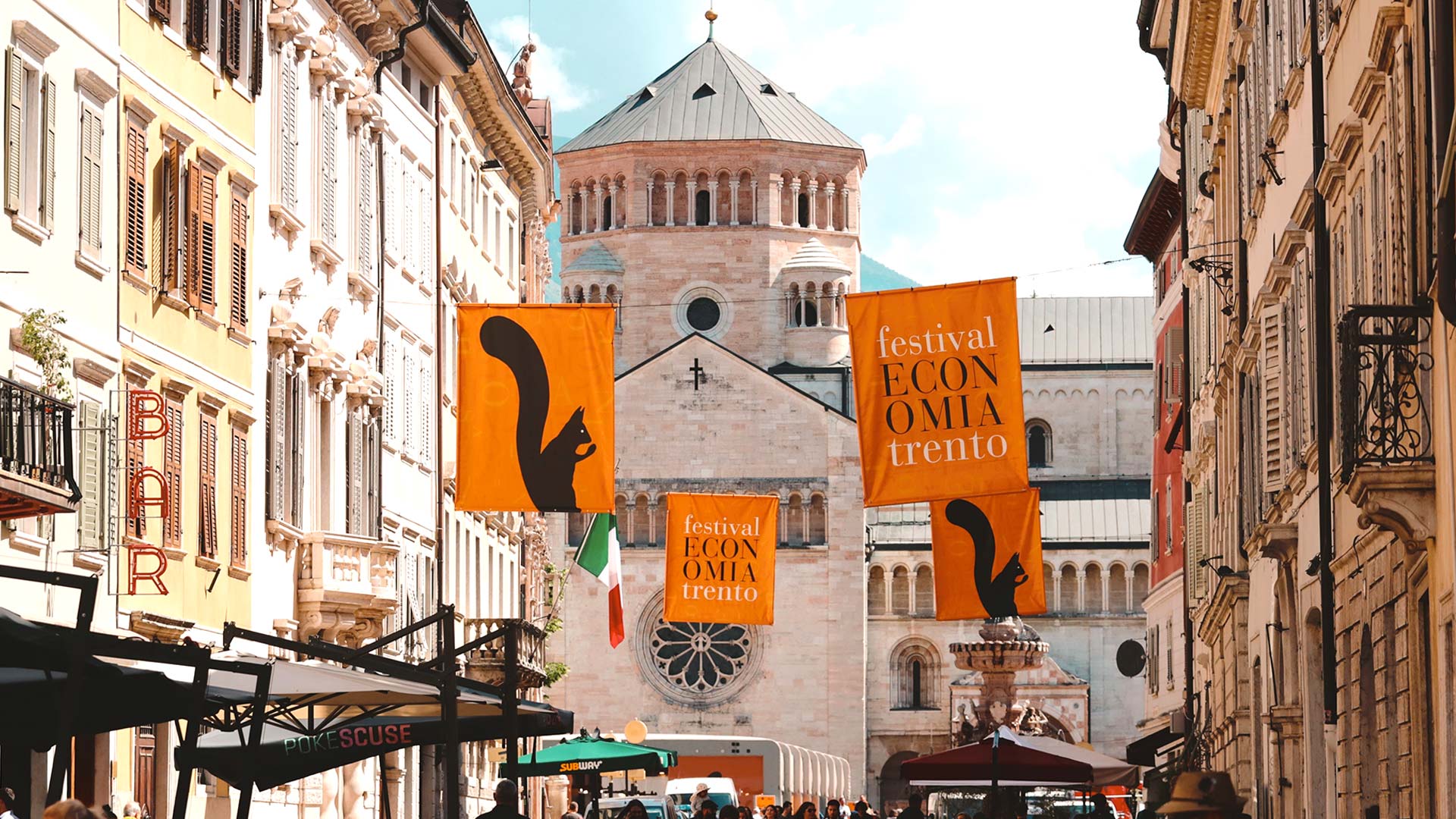 Bandiere del Festival dell'economia di Trento appese tra i palazzi del centro storico, con il Duomo di Trento sullo sfondo.